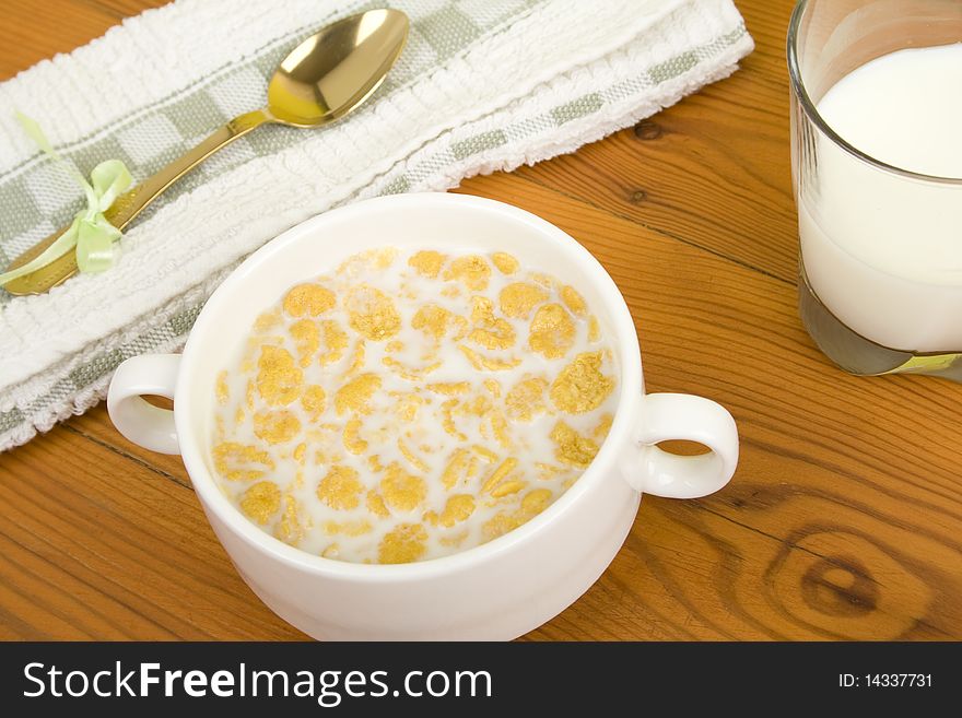 On the wooden table is breakfast cereal with milk, a glass of milk, a spoon, napkin, tea and flowers