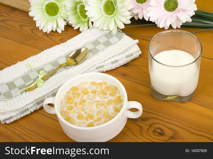 On the wooden table is breakfast cereal with milk, a glass of milk, a spoon, napkin, tea and flowers