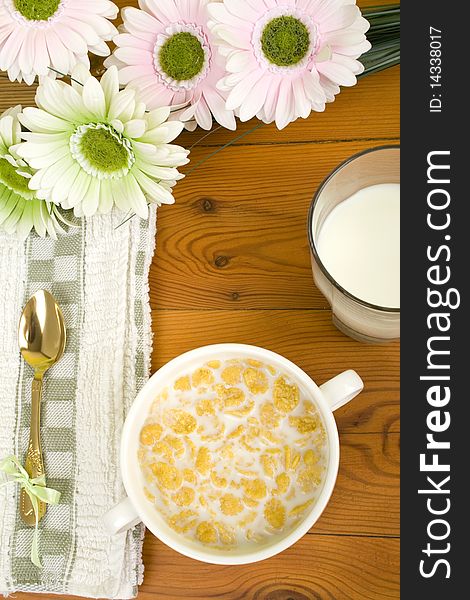 On the wooden table is breakfast cereal with milk, a glass of milk, a spoon, napkin, tea and flowers