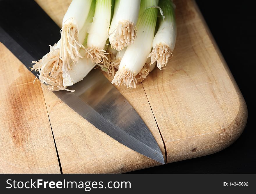 Fresh green onions on a wooden cutting board, with a knife, against a dark background. Fresh green onions on a wooden cutting board, with a knife, against a dark background
