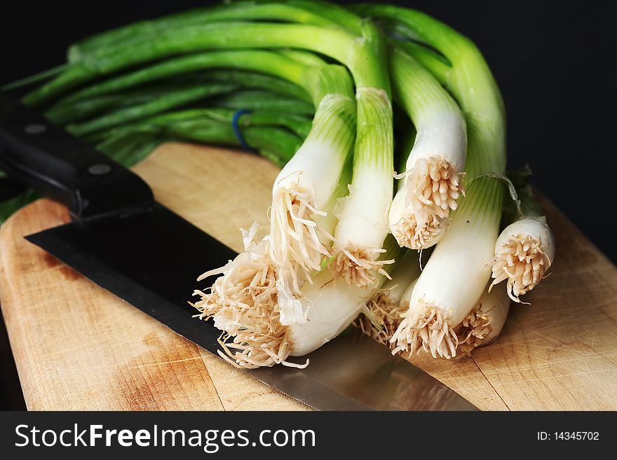 Fresh green onions on a wooden cutting board, with a knife, against a dark background. Fresh green onions on a wooden cutting board, with a knife, against a dark background