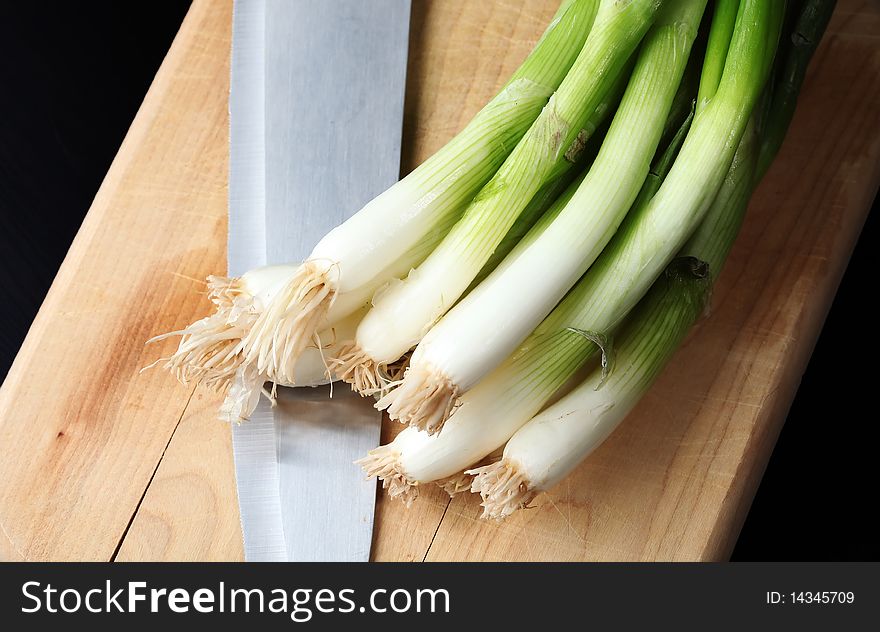 Fresh green onions on a wooden cutting board, with a knife, against a dark background. Fresh green onions on a wooden cutting board, with a knife, against a dark background