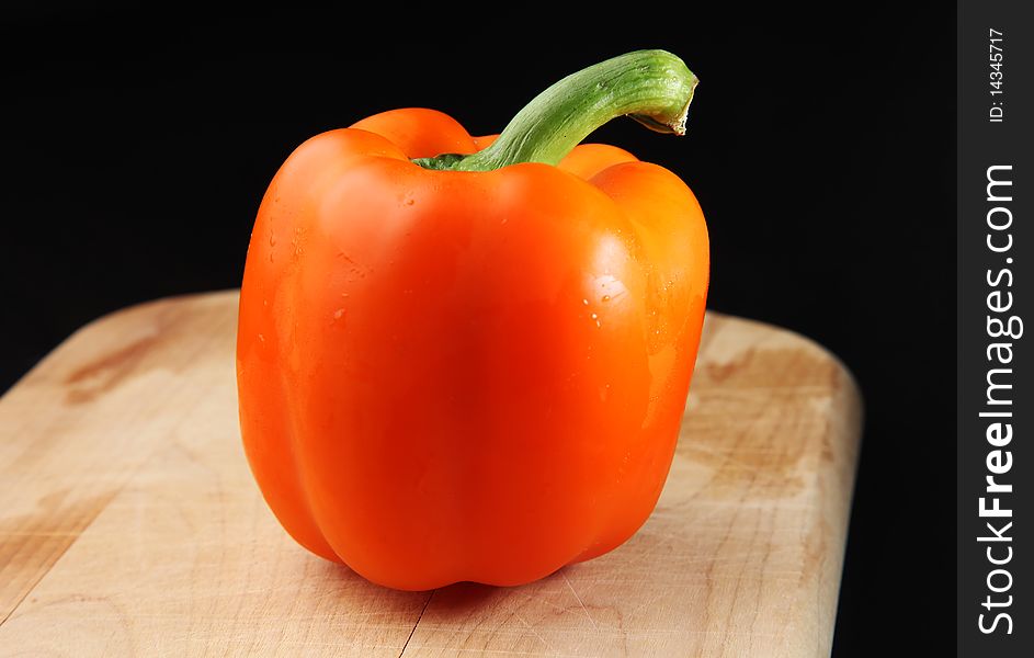 Fresh orange bell pepper, sitting on a wooden cutting board, against a dark background. Fresh orange bell pepper, sitting on a wooden cutting board, against a dark background.