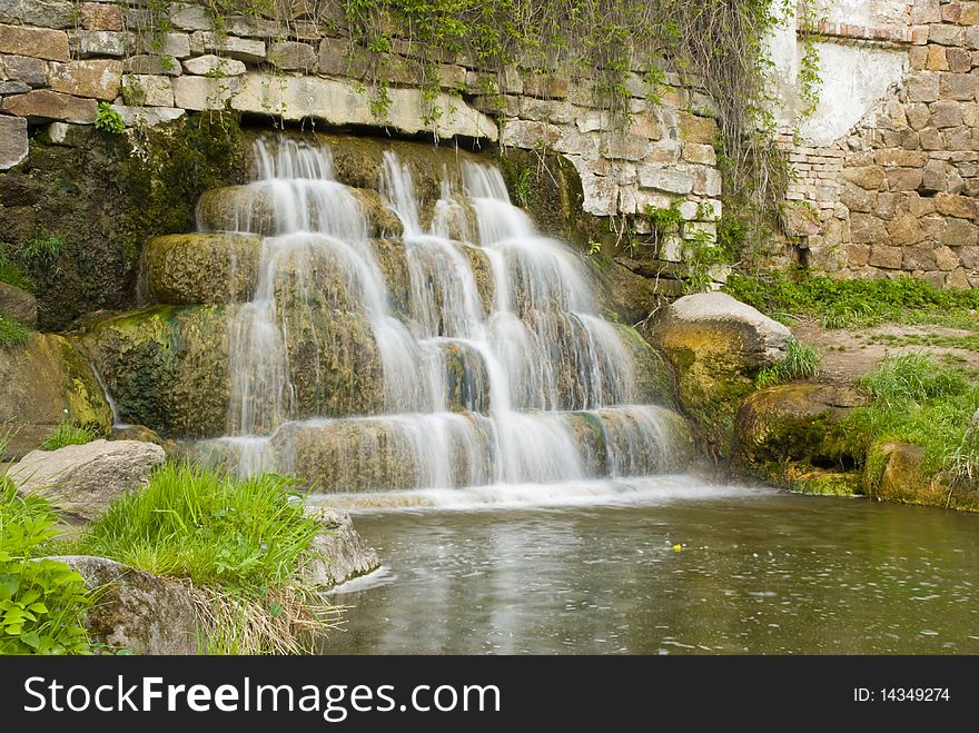 Waterfall flowing from the old wall and empties into the pond in the park on the shores of which lie boulders and juicy green grass grows. Waterfall flowing from the old wall and empties into the pond in the park on the shores of which lie boulders and juicy green grass grows