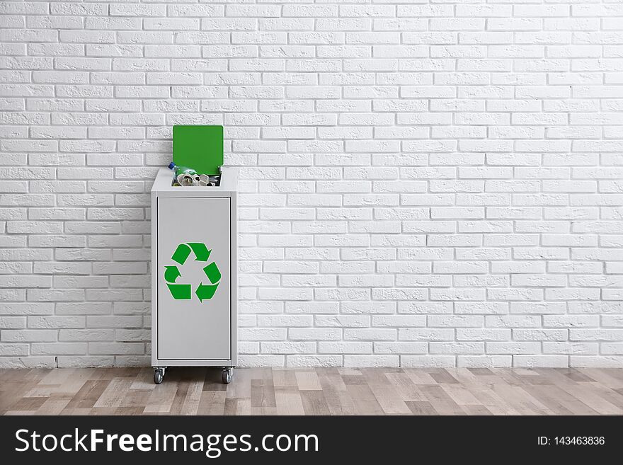 Overfilled trash bin with recycling symbol near brick wall indoors