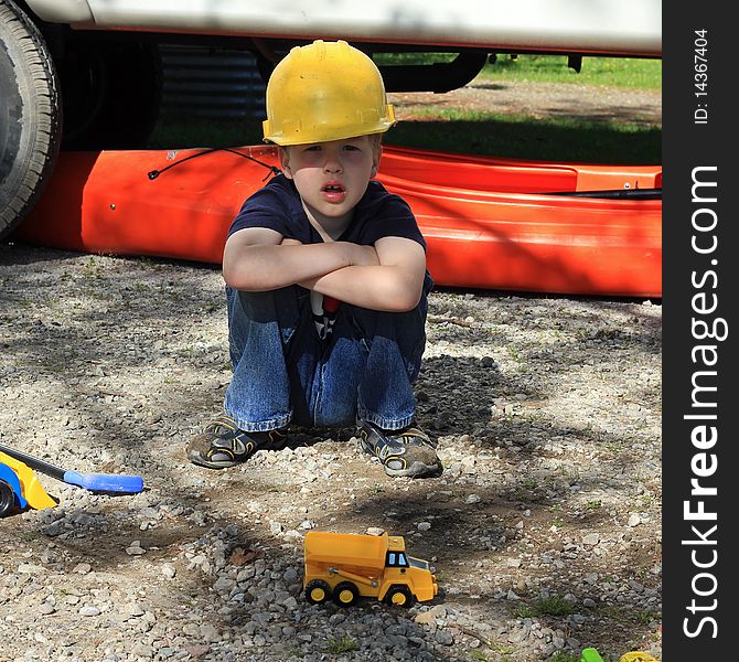 A young boy squatting on the ground wearing a yellow construction hard hat and playing with construction toys. He is at a campsite with an orange kayak in the background. A young boy squatting on the ground wearing a yellow construction hard hat and playing with construction toys. He is at a campsite with an orange kayak in the background.