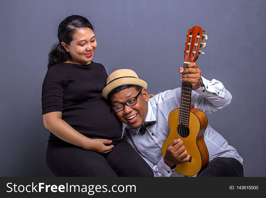 Happy couple expecting baby, men put his ear on wife& x27;s pregnant belly while playing music with an ukulele. Studio shot. Happy couple expecting baby, men put his ear on wife& x27;s pregnant belly while playing music with an ukulele. Studio shot