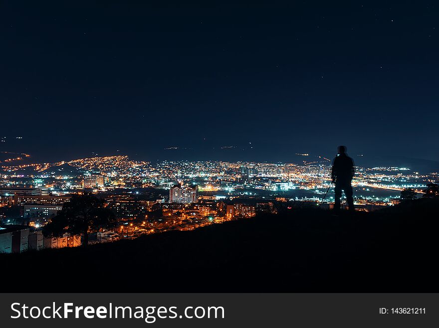 Night over big city. Photographer silhouette standing on top of the hill over the city, making night photography