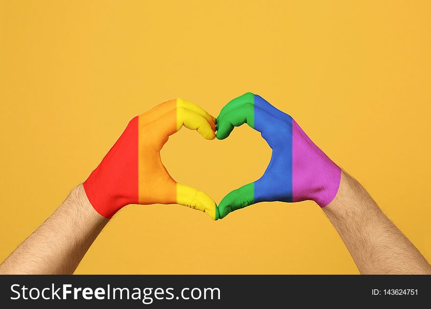 Man making heart with hands painted in LGBT flag on color background. Gay community