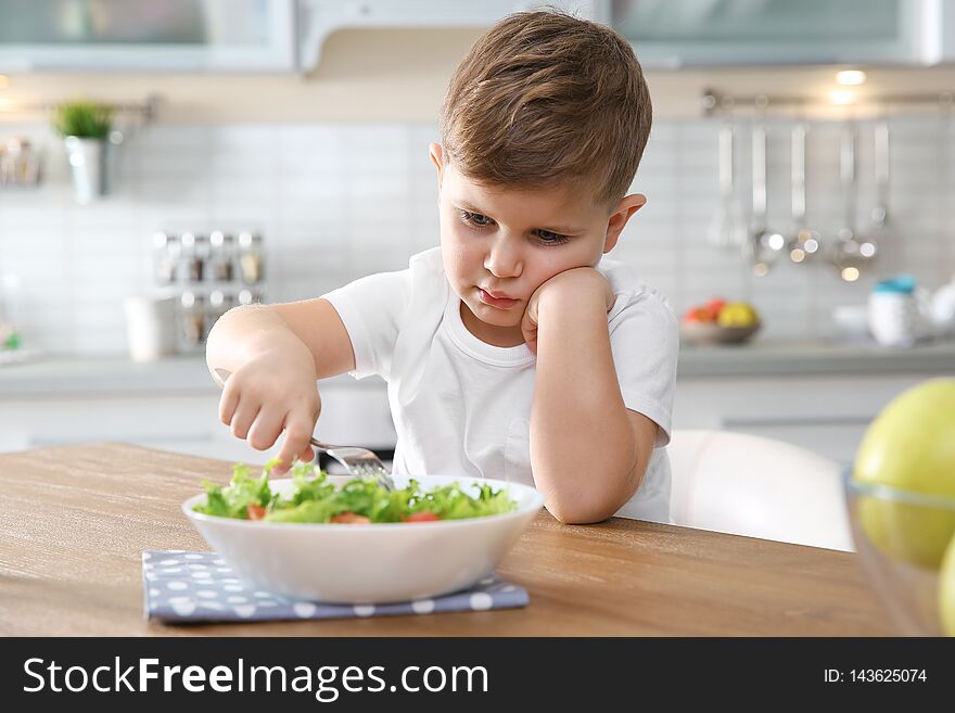 Unhappy little boy eating vegetable salad at table in kitchen