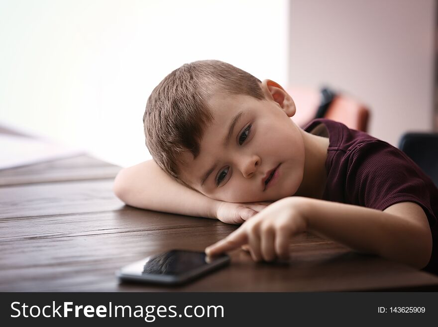 Sad little boy with mobile phone sitting at table indoors