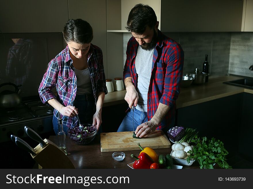 Beautiful young couple in kitchen at home while cooking healthy food. Husband cuts the cucumbers. Wife mix salad. Scene from