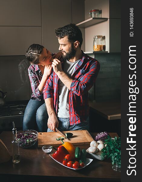 Beautiful young couple in kitchen at home while cooking healthy food. Husband cuts salad and feeds his wife cucumber. Scene from
