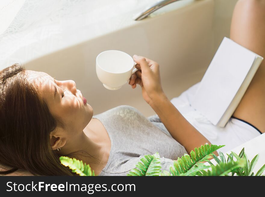 Beautiful Asian woman sleeps in a bathtub and holds a cup of coffee, morning with soft sunlight, with concept of relaxation and comfort on vacation