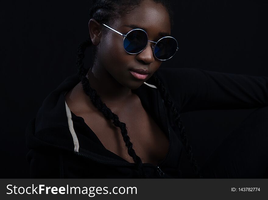 Portrait of a confident and cool young woman with dark skin wearing round sunglasses. High quality low-key studio portrait. Portrait of a confident and cool young woman with dark skin wearing round sunglasses. High quality low-key studio portrait.