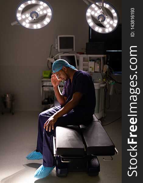 Side view of a nervous African-american male surgeon sitting with his head in his hand in operation room at hospital under a light ray. Side view of a nervous African-american male surgeon sitting with his head in his hand in operation room at hospital under a light ray