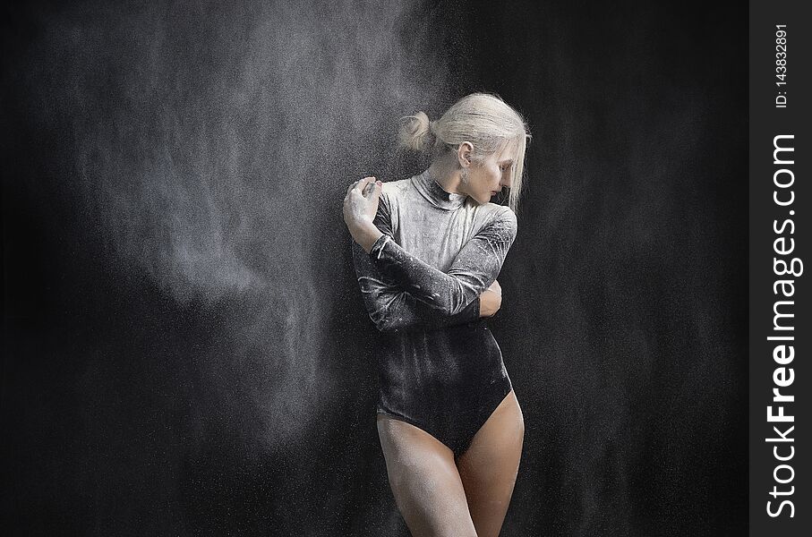 Beautiful slim girl wearing a black gymnastic bodysuit covered with clouds of the flying white powder dances on a dark background