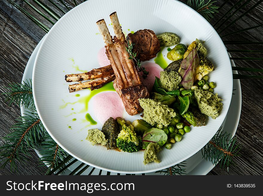 Top view closeup two pieces of grilled ribs with meat served with broccoli, artichokes, green peas and sauce on white restaurant plate on wooden table background. Top view closeup two pieces of grilled ribs with meat served with broccoli, artichokes, green peas and sauce on white restaurant plate on wooden table background