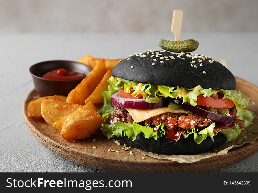 Wooden plate with black burger and french fries on table, closeup