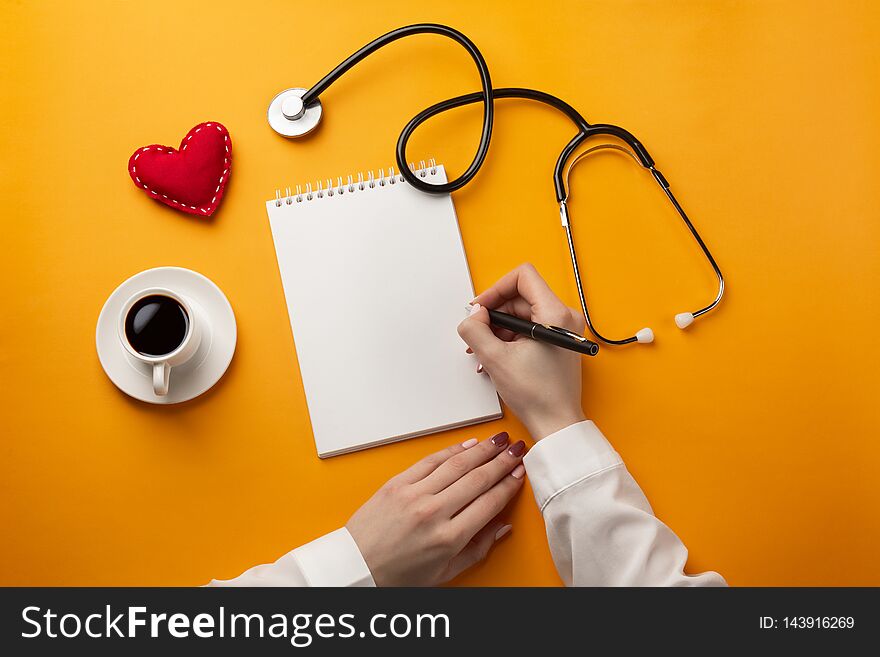 Professional doctor writing medical records in a notebook with stethoscope, coffee cup, syringe and heart. Top view with place for your text