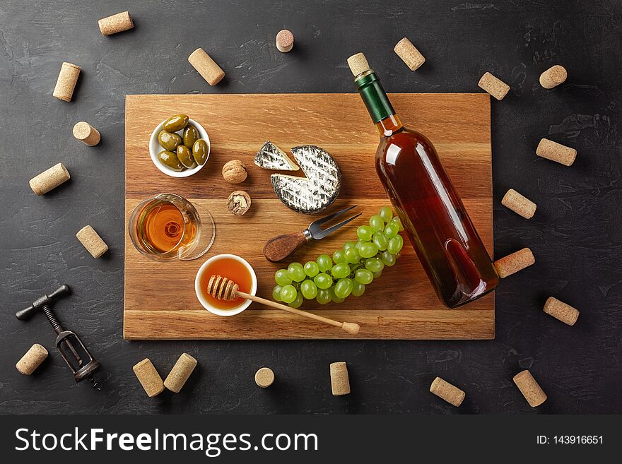 White wine bottle, cheese head, bunch of grapes, honey, nuts and wineglass on cutting board with corks and corkscrew on black