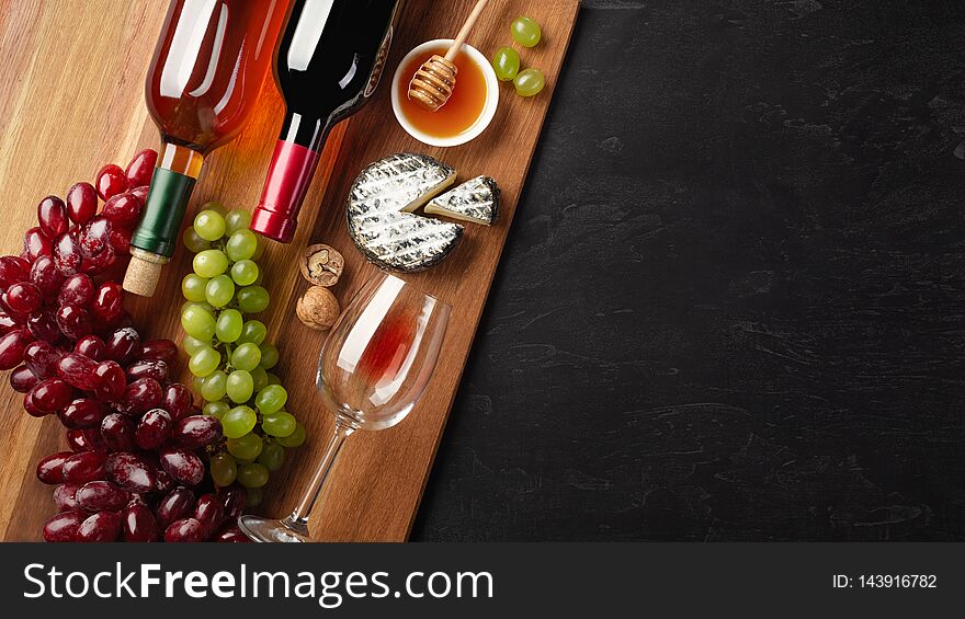 Red and white wine bottles with bunch of grapes, cheese, honey, nuts and wineglass on wooden board and black background. Top view with copy space.