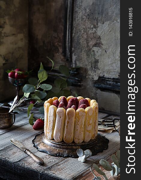 Homemade traditional Italian cake tiramisu with cookies around the cake and strawberries on the top on silver tray on wooden table with vintage cups, knife, leaves opposite concrete wall