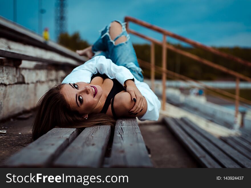 Young Sexy Woman Lies On A Wooden Bench. She Is Taking Break After Workout In Gym. Outdoor