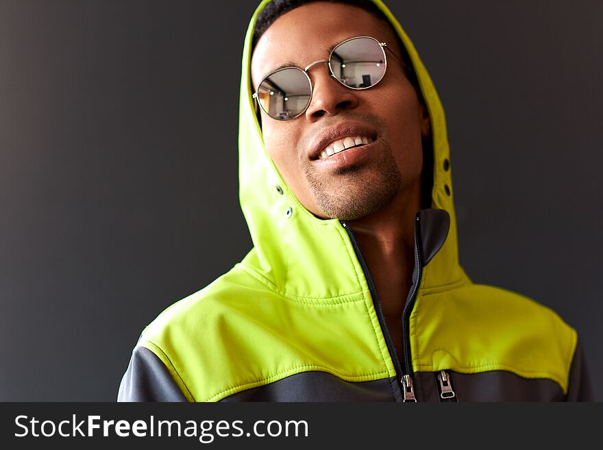 Stylish attractive African American man smiling and looking through mirror sunglasses, wearing green sport hoody, posing on black studio wall. People, emotion and lifestyle concept