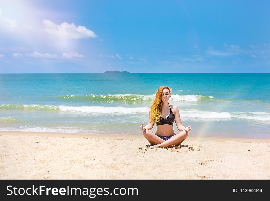 A young, beautiful girl with red hair, in a bikini. practices yoga on the white sand and turquoise sea.