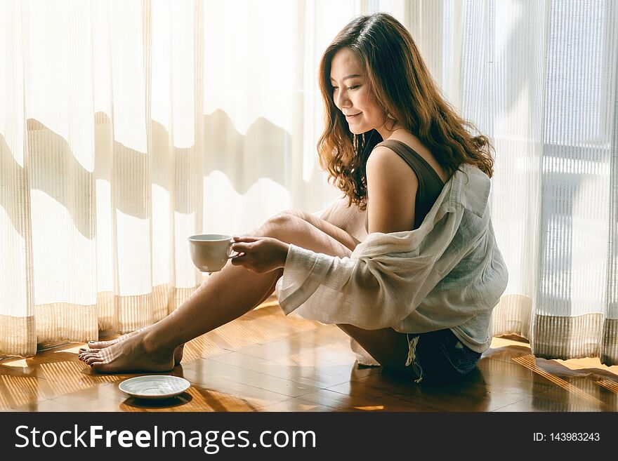 A beautiful asian woman sitting and holding a cup of hot coffee to drink on the floor in the morning