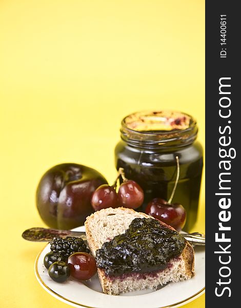 Blackberry jam in an open jar and on one piece of sliced bread on white plate against yellow background. Plum, cherries, blackberries and blueberries surround the jam. Blackberry jam in an open jar and on one piece of sliced bread on white plate against yellow background. Plum, cherries, blackberries and blueberries surround the jam.