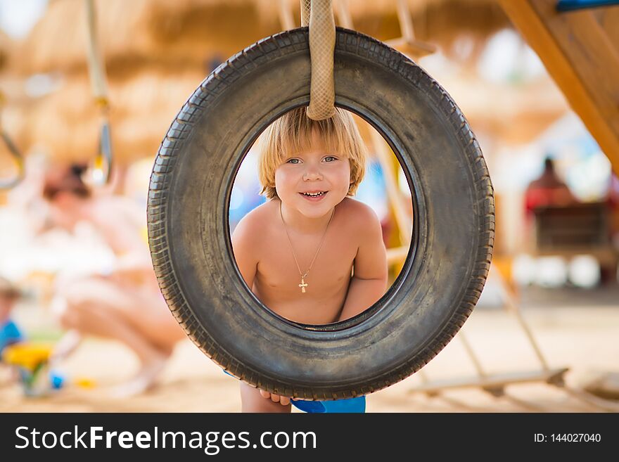 Cute, excited, blond-haired child boy playfully looking through the tire swing on the modern playground near the beach. Cute, excited, blond-haired child boy playfully looking through the tire swing on the modern playground near the beach