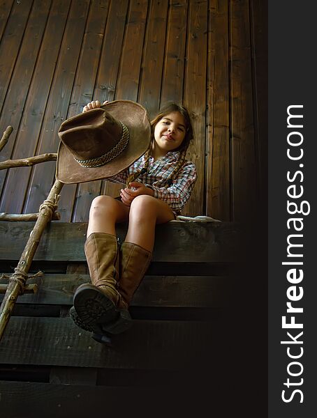 Little girl in a wide-brimmed cowboy hat and traditional dress in high boots and with a lasso posing on a dark wooden background. Little girl in a wide-brimmed cowboy hat and traditional dress in high boots and with a lasso posing on a dark wooden background