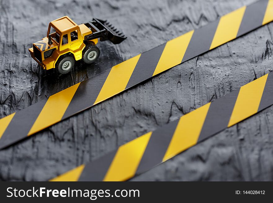 Yellow Toy Bulldozer On A Black Background Texture. Black And Yellow Fence Tape Against The Background Of The CONCEPT