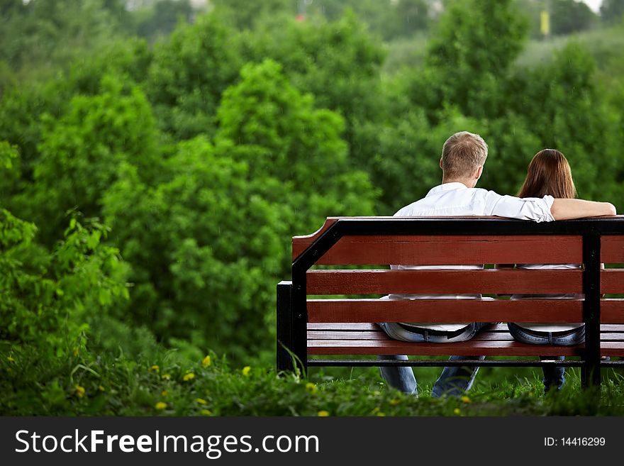 The enamoured couple sits on a bench against green trees