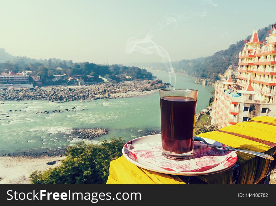 A glass of coffee on a plate against the background of the beautiful scenery of Rishikesh.. View of Ganga river embankment, Lakshman Jhula bridge and Tera Manzil Temple, Trimbakeshwar in Rishikesh