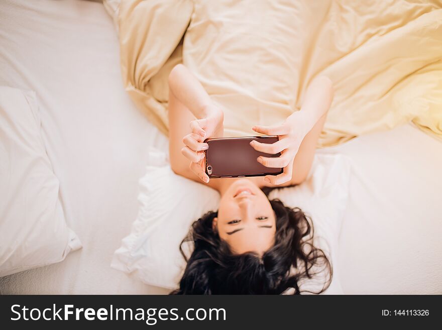 Beautiful girl with black wavy hair naked taking a selfie on phone lying in the morning in bed. Indoor shot of cheerful caucasian woman has morning video call, holds modern cell phone. Beautiful girl with black wavy hair naked taking a selfie on phone lying in the morning in bed. Indoor shot of cheerful caucasian woman has morning video call, holds modern cell phone