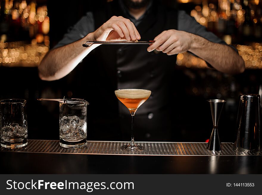 Male bartender adding to a cocktail in the glass a grated nutmeg on the bar counter in the dark blurred background