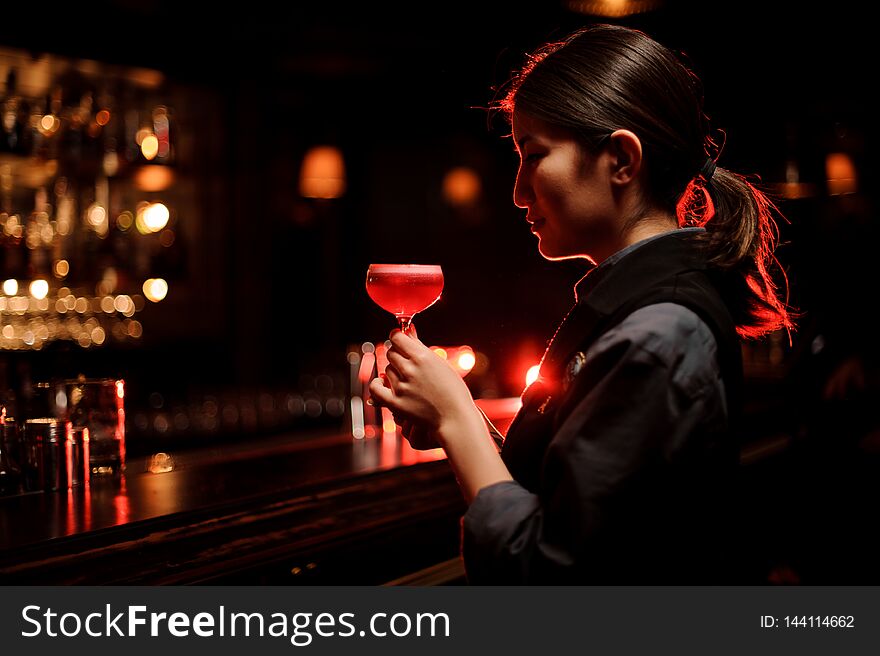 Bartender girl holding a smooth crimson cocktail in the glass with a one pink rose bud as a decor
