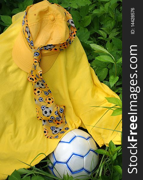 Yellow baseball cap, t-shirt, tie with soccer balls on it and soccer ball against green leaves. Yellow baseball cap, t-shirt, tie with soccer balls on it and soccer ball against green leaves.