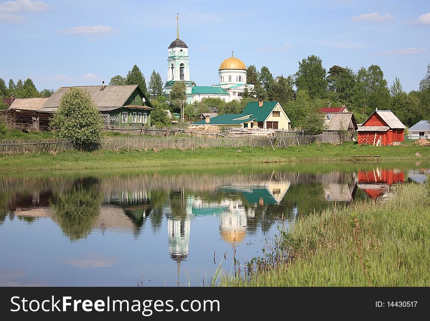 The image of an old orthodox church, rural wooden houses reflecting in the calm water of a river. The image of an old orthodox church, rural wooden houses reflecting in the calm water of a river