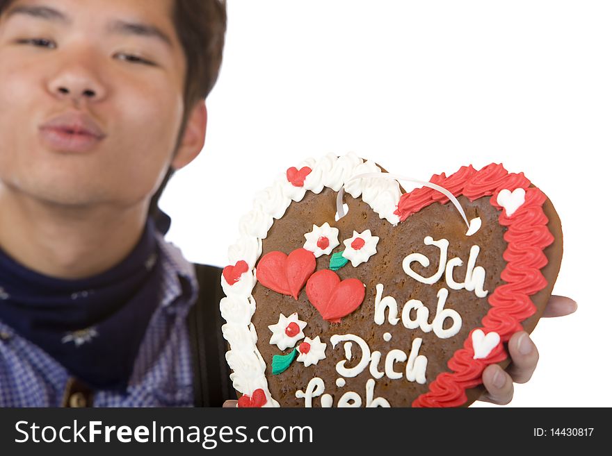 Asian man, dressed with Bavarian Lederhose holds an Oktoberfest gingerbread heart in camera and gives a kiss. Isolated on white. Asian man, dressed with Bavarian Lederhose holds an Oktoberfest gingerbread heart in camera and gives a kiss. Isolated on white.
