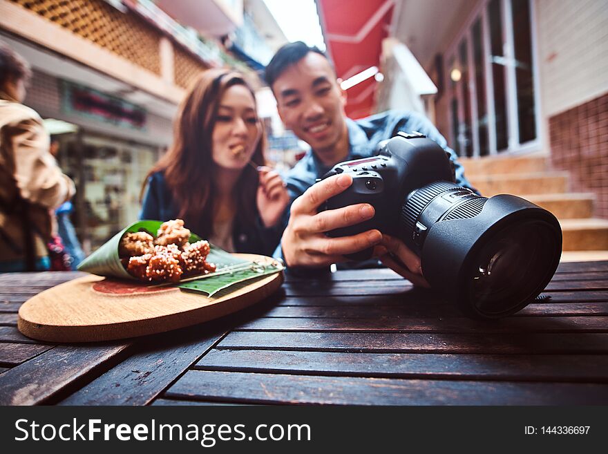 Happy young chinese couple sitting at cafe outside enjoying traditional asian food and watching pictures in photo camera. Widescreen shooting. Happy young chinese couple sitting at cafe outside enjoying traditional asian food and watching pictures in photo camera. Widescreen shooting.