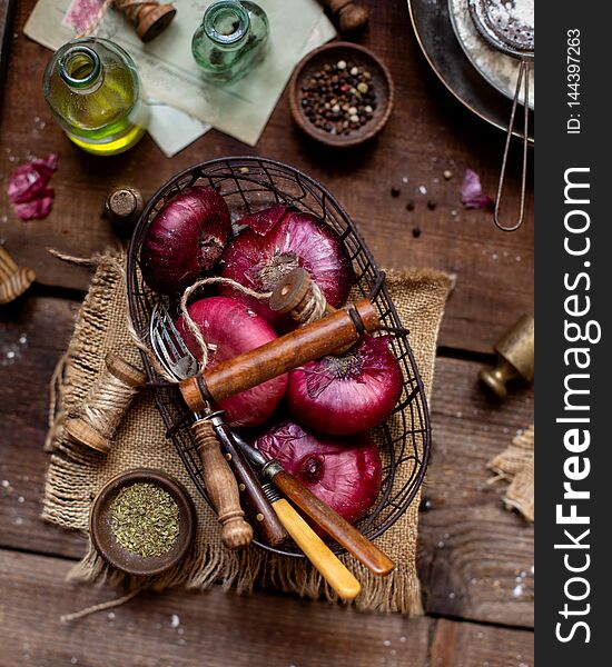 Overhead shot of metal basket of red onion, spoons, strings standing on wooden brown table with sackcloth, bottle with oil, spices, pepper, plate with flour, vintage weights. Ingredients for cooking. Overhead shot of metal basket of red onion, spoons, strings standing on wooden brown table with sackcloth, bottle with oil, spices, pepper, plate with flour, vintage weights. Ingredients for cooking