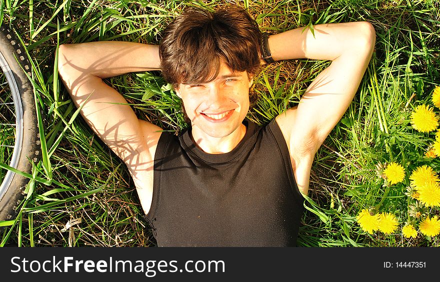 Portrait of a happy young man lying on the grass. Bike wheel is present on the left, a few dandelions on the right; warm lighting setup. Portrait of a happy young man lying on the grass. Bike wheel is present on the left, a few dandelions on the right; warm lighting setup