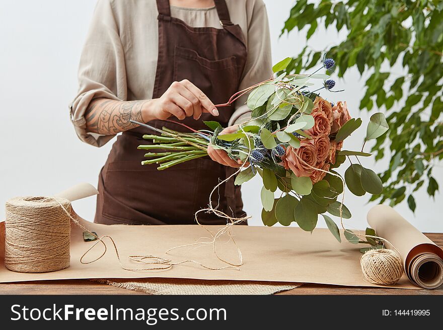 Female florist is decorating beautiful bouquet from fresh natural roses step by step at the table with paper and rope on