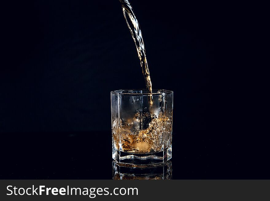 Whiskey pouring into glass with ice isolated on reflective black surface. Whiskey splash out of glass, many drops of beverage get out from glass. Whiskey pouring into glass with ice isolated on reflective black surface. Whiskey splash out of glass, many drops of beverage get out from glass