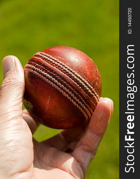 Used red leather cricket ball being held by hand against a green grass background