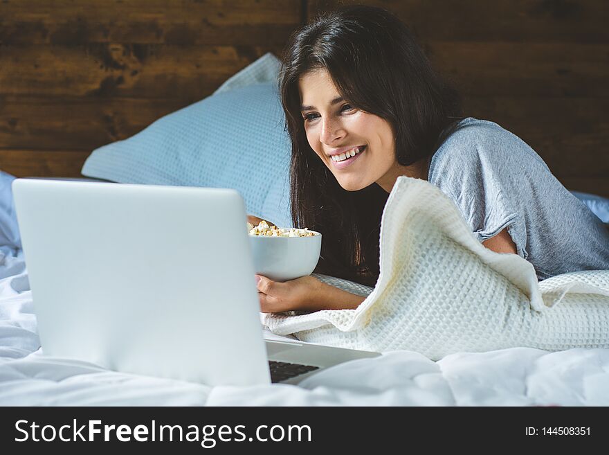 Close-up of young woman using laptop and eating popcorn in a comfortable bed at home